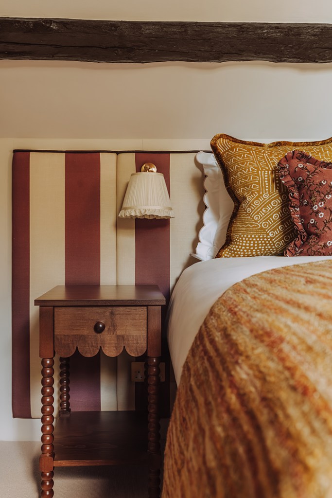 A close-up view of a stylish bedroom featuring a wooden bedside table with a drawer, a decorative lamp, and patterned cushions on the bed against a striped headboard.