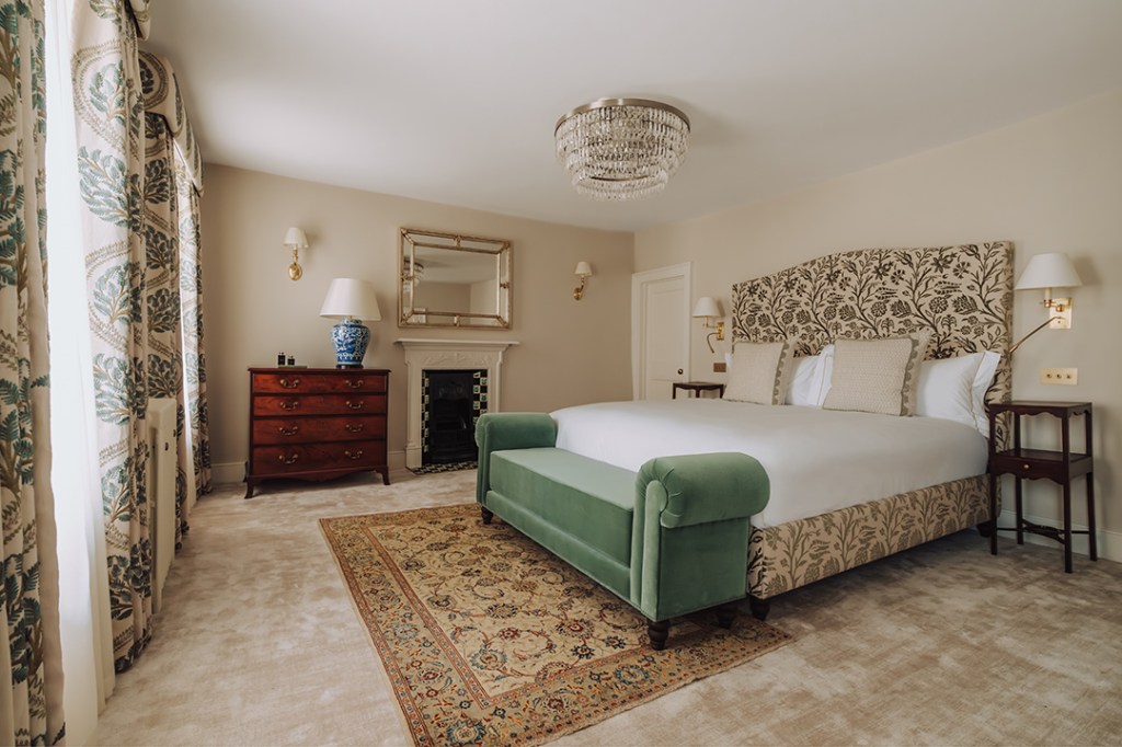 Luxurious hotel bedroom featuring a large bed with a floral headboard, green upholstered end bench, vintage wooden dresser, and decorative curtains. A chandelier hangs from the ceiling, and a patterned rug adds warmth to the carpeted floor.