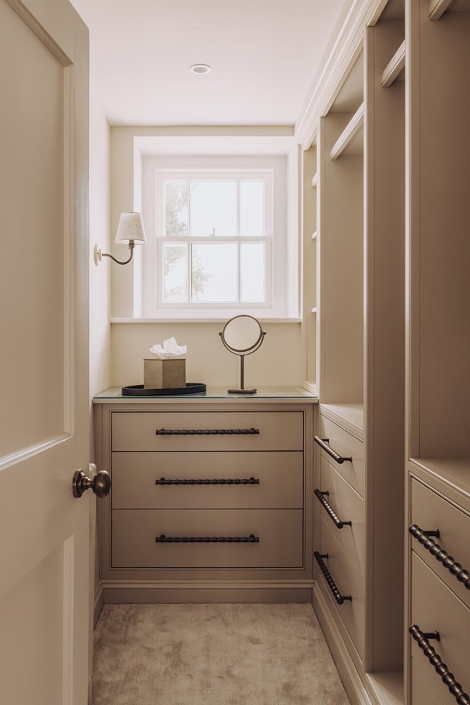 A well-organised dressing room with a window, featuring a chest of drawers, a mirror, and soft lighting.