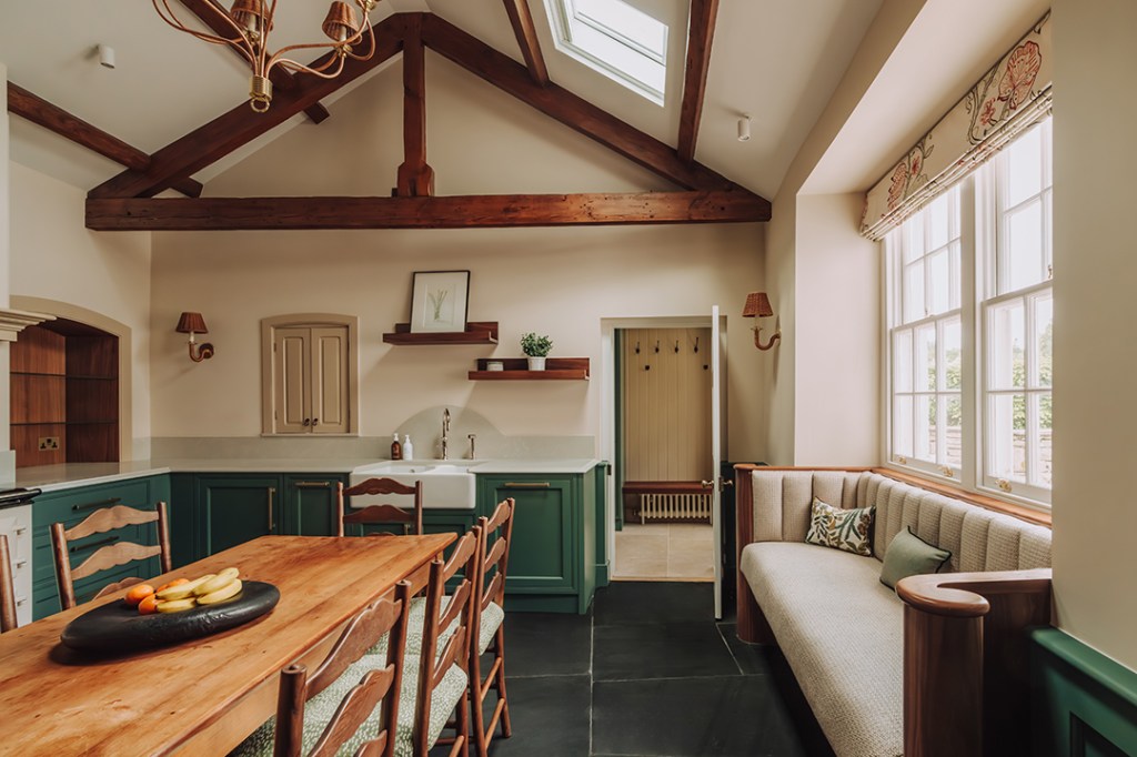A modern kitchen featuring a wooden dining table with fruit, green cabinetry, a white sink, and large windows with natural light. Exposed wooden beams and a comfortable seating area add to the design.