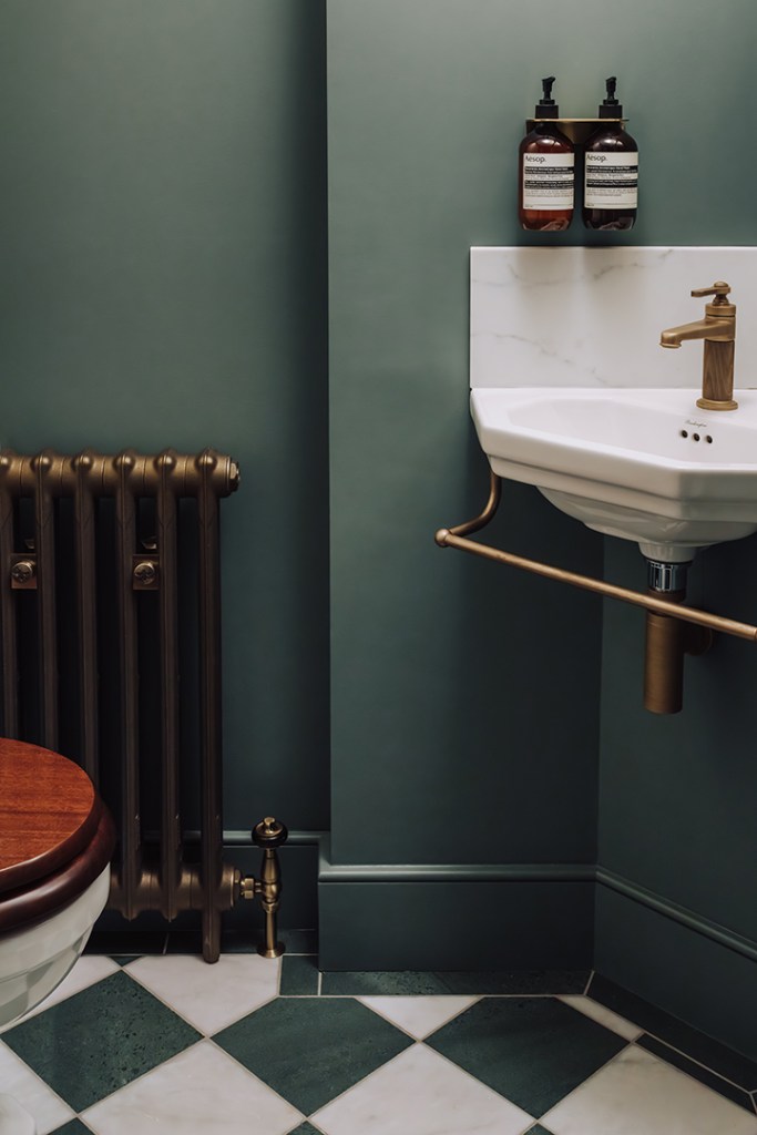 A stylish bathroom corner featuring a white sink, a gold tap, and dark green walls. There is a vintage radiator and two bottles of Aesop products displayed above the sink.