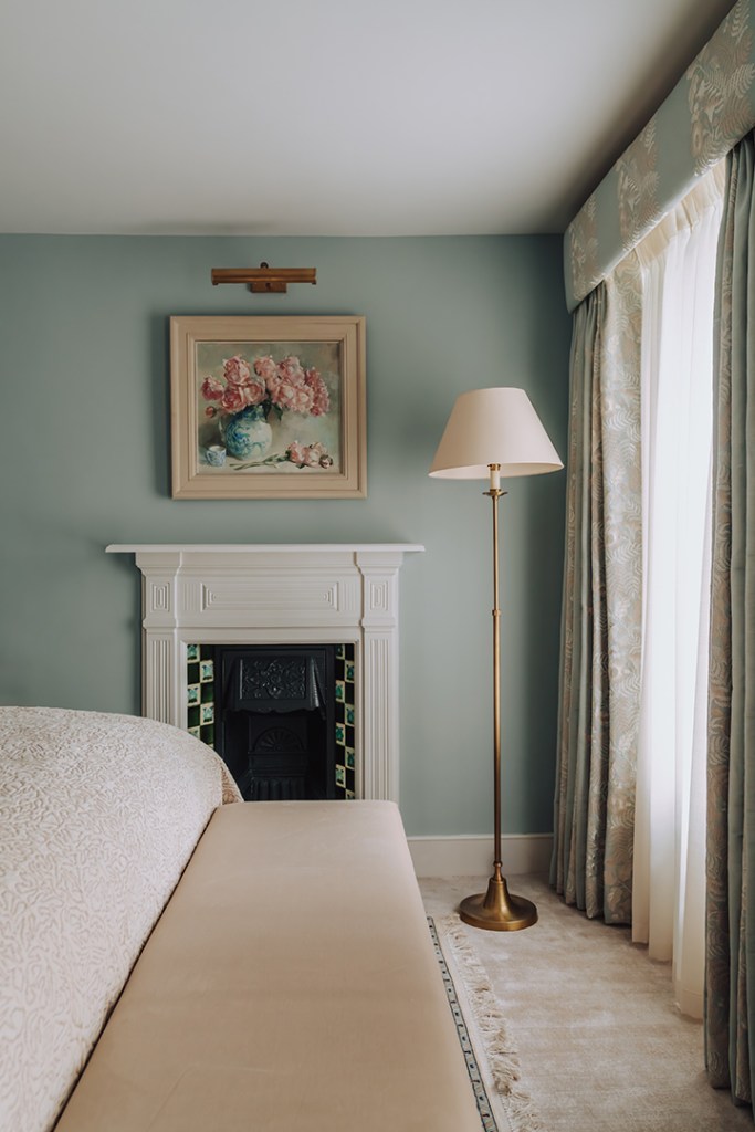 A cosy bedroom featuring a beige bed, a floral painting on the wall, a floor lamp with a cream shade, and a white fireplace with decorative tiles, all set against a pale blue wall.