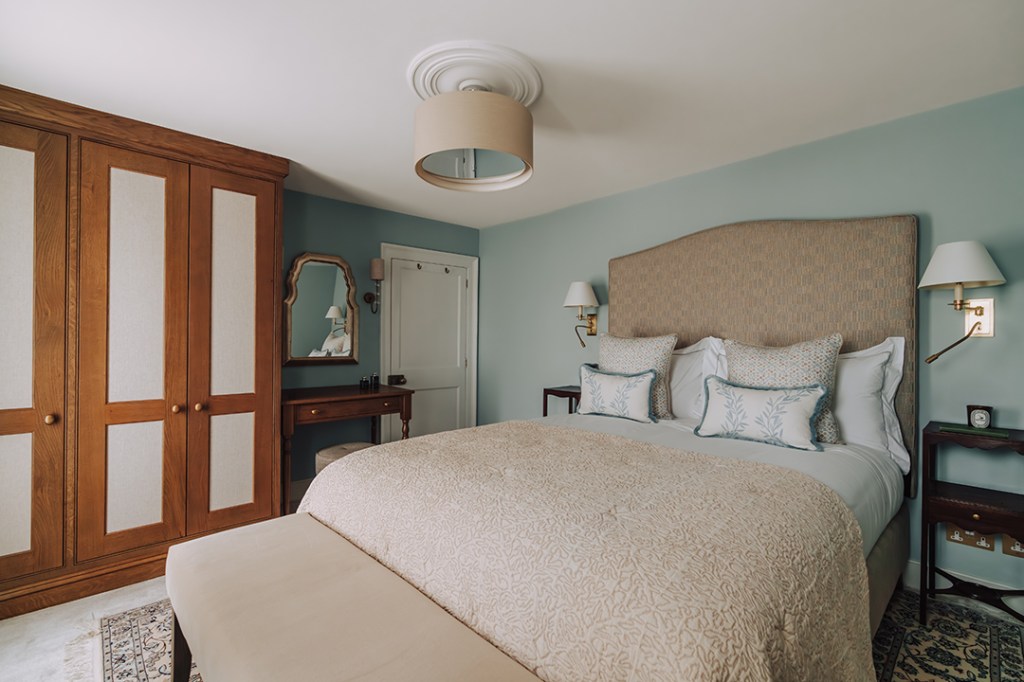 A stylish bedroom featuring a large bed with decorative pillows, a beige bedspread, and a padded headboard. To the left, there is a wooden wardrobe with patterned doors, and a small dressing table with a mirror. The walls are painted a soft blue, and there are bedside lamps beside the bed, creating a cozy atmosphere.
