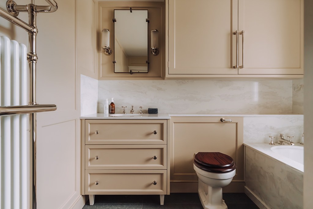 Elegant bathroom featuring a beige vanity unit with drawers, a large mirror, and a marble splashback. Includes a traditional toilet with a wooden seat and a bathtub alongside.