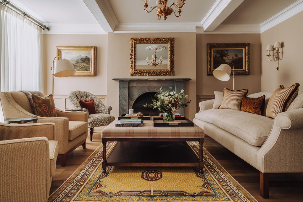 A stylish living room featuring a cream sofa and armchairs, a coffee table with books, and a marble fireplace. Decorative paintings and a chandelier enhance the elegant atmosphere.